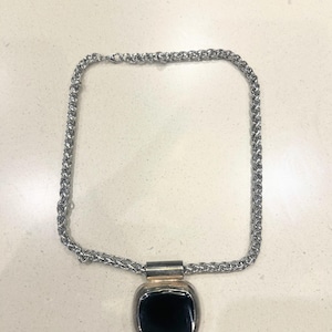 May include: Silver necklace featuring a square black pendant. The chain has a woven pattern, and the pendant is a dark, glossy square set in a silver frame. The necklace is shown on a light-coloured surface.