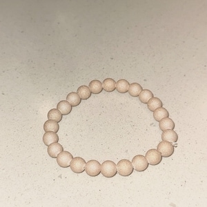 May include: A bracelet made of light beige, spherical beads. The beads are strung together to form a circular shape. The beads have a textured surface and are likely made from a natural material. The bracelet is elasticated.