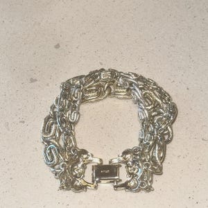 May include: Silver-toned bracelet with a detailed chain link design. The bracelet features an ornate clasp with a rectangular shape. The links are intricately woven, creating a textured appearance. The jewelry is displayed on a neutral surface.