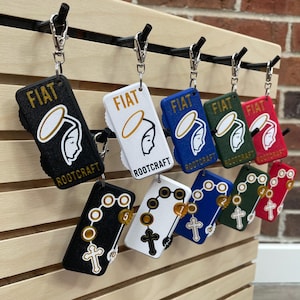 May include: A selection of keyrings with religious imagery. Each keyring has a rectangular shape, displaying the word "FIAT" and a depiction of a figure with a halo. The keyrings are available in multiple colours, such as black, white, blue, green, and red.