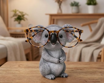 May include: A gray koala figurine designed as an eyeglass holder, featuring a textured fur appearance, a black nose, and round eyes. The koala is wearing a pair of brown and black tortoiseshell patterned eyeglasses resting on its face.