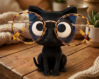 May include: A matte black cat-shaped eyeglass holder featuring large, wide-eyed facial features. The figurine is wearing a pair of round, tortoiseshell-patterned glasses with thin gold-toned metal frames resting across its nose and eyes.