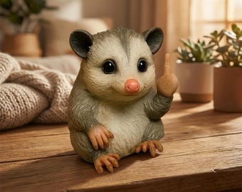 May include: A whimsical figurine of an opossum giving the middle finger. The opossum is light gray and tan with a pink nose and black eyes. It is sitting on a wooden surface. The figurine is a humorous decorative item.