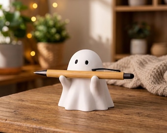 May include: A white ceramic ghost-shaped pen holder with black dot eyes, holding a wooden pen. The pen has a black tip and clip. The holder sits on a wooden surface, with plants and a shelf in the background.