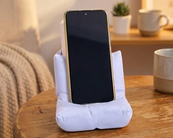 May include: A smartphone with a black screen and a gold-colored frame resting in a light lavender, soft, padded, rectangular phone stand. The stand features a plush, pillow-like design that supports the device in an upright, vertical position.