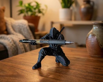 May include: A matte black 3D-printed ninja figurine in a kneeling position, holding a Pilot G-2 07 black ink gel pen horizontally with both hands. The figure features detailed fabric folds on the clothing and a sword hilt visible on the back.