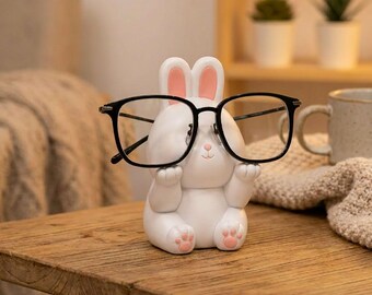 May include: A white rabbit figurine with pink inner ears and pink paw pads, designed as a stand for eyewear. The rabbit is in a seated position with its front paws raised, holding a pair of black-framed glasses across its face.