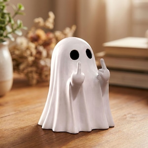 Può includere: Statuetta a forma di fantasma in ceramica bianca con fori neri per gli occhi e dita medie alzate. Il fantasma è drappeggiato in una forma a lenzuolo con un fondo a balze. La statuetta è su una superficie di legno.