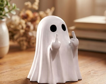Puede incluir: Figura de fantasma de cerámica blanca con agujeros negros para los ojos y dedos medios levantados. El fantasma está envuelto en una forma de sábana con un fondo con volantes. La figura está sobre una superficie de madera.