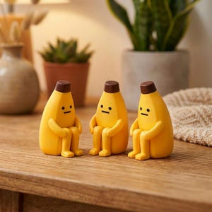 Könnte beinhalten: Drei gelbe, bananenförmige Figuren mit braunen Oberteilen und einfachen Gesichtszügen. Die Figuren sitzen mit verschränkten Armen und erzeugen eine skurrile und verspielte Ästhetik. Sie werden auf einer Holzoberfläche präsentiert.