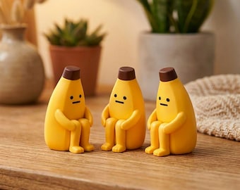 May include: Three yellow banana-shaped figurines with brown tops and simple facial features. The figurines are seated with their arms crossed, creating a whimsical and playful aesthetic. They are displayed on a wooden surface.