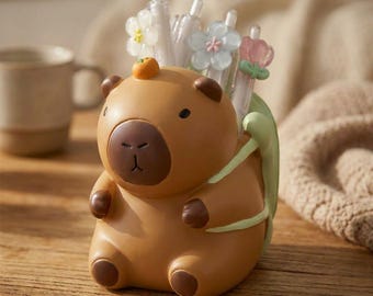 May include: A brown bear-shaped desk organizer with a small orange fruit on its head. The organizer holds pens and pencils with flower-shaped toppers. The bear has a brown nose and ears, and green leaves are on its back.