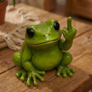 May include: A green frog ornament with large black eyes and a raised middle finger. The frog is positioned on a wooden surface. The ornament is a humorous decorative item.