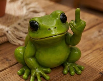 May include: A green frog figurine with large black eyes and a raised middle finger. The frog is sitting on a wooden surface. The figurine is a humorous decorative item.