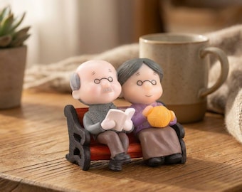 May include: A miniature figurine of an elderly couple sitting on a small red and black bench. The figure on the left is a bald person in a gray sweater reading a white book. The figure on the right has gray hair, wears a purple top, and holds a yellow ball of yarn.