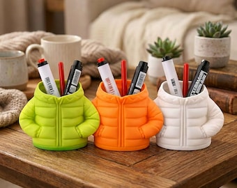 May include: Three desk organizers shaped like miniature puffer jackets in lime green, bright orange, and white. Each organizer features a quilted texture, a central zipper detail, and two side pockets, holding various markers and pens upright.