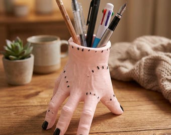May include: A pink hand-shaped pen holder with black stitched details and black fingernails. The holder contains a pencil, pens, and a fountain pen. The holder is on a wooden surface.
