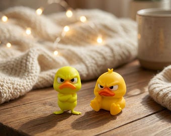 May include: Two small yellow duck figurines with orange beaks and angry facial expressions. One duck stands with its wings crossed over its chest, while the other sits with its legs extended forward, both featuring large, furrowed, dark eyebrows.