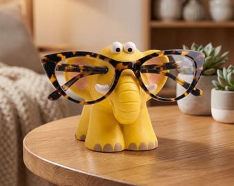 May include: A yellow elephant-shaped eyeglass holder with two white eyes and small tusks. The elephant is holding a pair of brown and black tortoiseshell-patterned cat-eye glasses across its trunk and ears, providing a stable stand for eyewear.