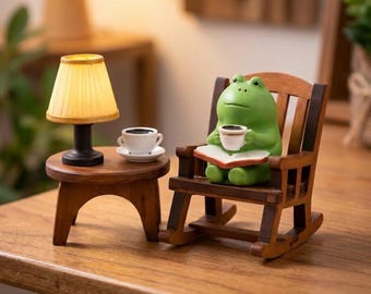 May include: A whimsical miniature scene featuring a green frog figurine seated in a wooden rocking chair, holding a cup of coffee and reading a book. A small wooden side table holds a lamp and a cup of coffee. The lamp has a yellow lampshade.