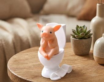 May include: A small, light pink pig figurine sitting on a white toilet. The pig has large ears and a rounded snout. A small, white, coiled roll of toilet paper is attached to the base of the white toilet, which has a smooth, glossy finish.