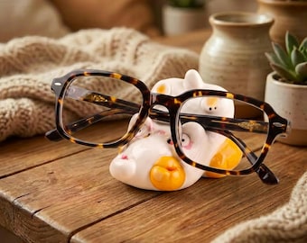 May include: A pair of tortoise shell patterned eyeglasses resting on a white ceramic animal-shaped holder. The holder features rounded ears, small pink accents, and bright yellow circular details. The glasses have a classic rectangular frame.