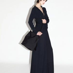 Pode incluir: Uma mulher vestindo um longo vestido azul marinho escuro com mangas compridas e decote em V. O vestido é combinado com uma bolsa de ombro preta. A roupa é simples e elegante, adequada para ocasiões formais ou uso diário.