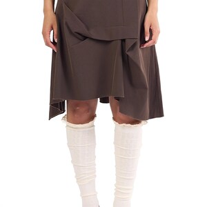 Op de afbeelding: Een donkerbruine knielange rok met een asymmetrische zoom en gestructureerde plooien in de taille. De rok wordt gecombineerd met gebroken witte geribbelde kniekousen met een fijn kanten randje aan de bovenkant en zwarte gesloten schoenen.