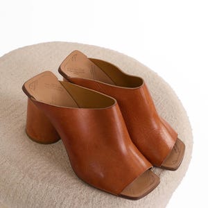 May include: A pair of tan leather mules featuring an open-toe design, a square toe box, and a thick, cylindrical block heel. The interior footbed is lined in a lighter tan leather and displays visible brand text and size markings.