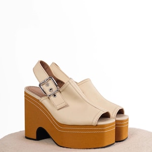 May include: A pair of cream-coloured open-toe platform sandals featuring a thick mustard-yellow wedge sole with visible white stitching. The sandals include a slingback strap secured by a silver-toned metal buckle on the side of the shoe.