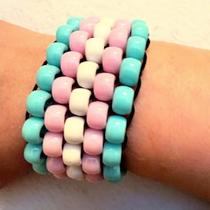 May include: A multi-row bracelet constructed from smooth, cylindrical plastic pony beads. The beads are arranged in a repeating pattern of light blue, light pink, and white, strung together on thin black elastic cords to form a wide band.