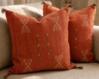 May include: Two square throw pillows in a deep burnt orange color featuring a textured fabric weave. Each pillow is adorned with small, light-colored geometric patterns and finished with tiny tassels at each of the four corners of the covers.