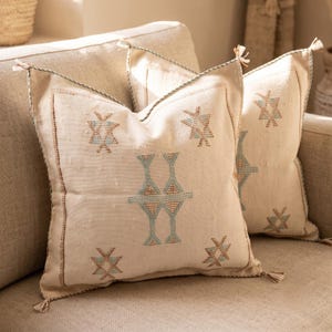 May include: Two square off-white throw cushions featuring embroidered geometric patterns in light blue and brown. Each cushion has a decorative border and small tassels at the four corners, crafted from a textured fabric with a visible weave.