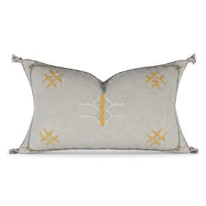 Puede incluir: Cojín decorativo rectangular con fondo gris claro y patrones geométricos bordados en amarillo y blanco. El cojín tiene borlas en cada esquina y un borde gris oscuro.