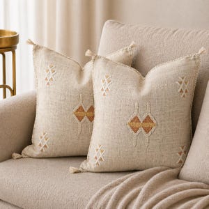 May include: Two beige decorative throw cushions with geometric embroidery in white and gold. The cushions have a textured appearance and are adorned with tassels at each corner. They are placed on a beige sofa.