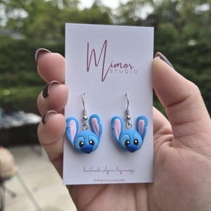 Pu&ograve; includere: Un paio di orecchini in pasta polimerica blu e rosa a forma di personaggio dei cartoni animati. Gli orecchini sono esposti su una carta bianca con la scritta "Mimor Studio" e "handmade polymer clay earrings".