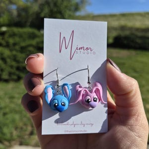 Puede incluir: Pendientes hechos a mano de arcilla polim&eacute;rica, que representan un personaje de dibujos animados azul y otro rosa con orejas grandes. Los pendientes se muestran en una tarjeta blanca con el logotipo "Mimor Studio" y texto. Fondo borroso.