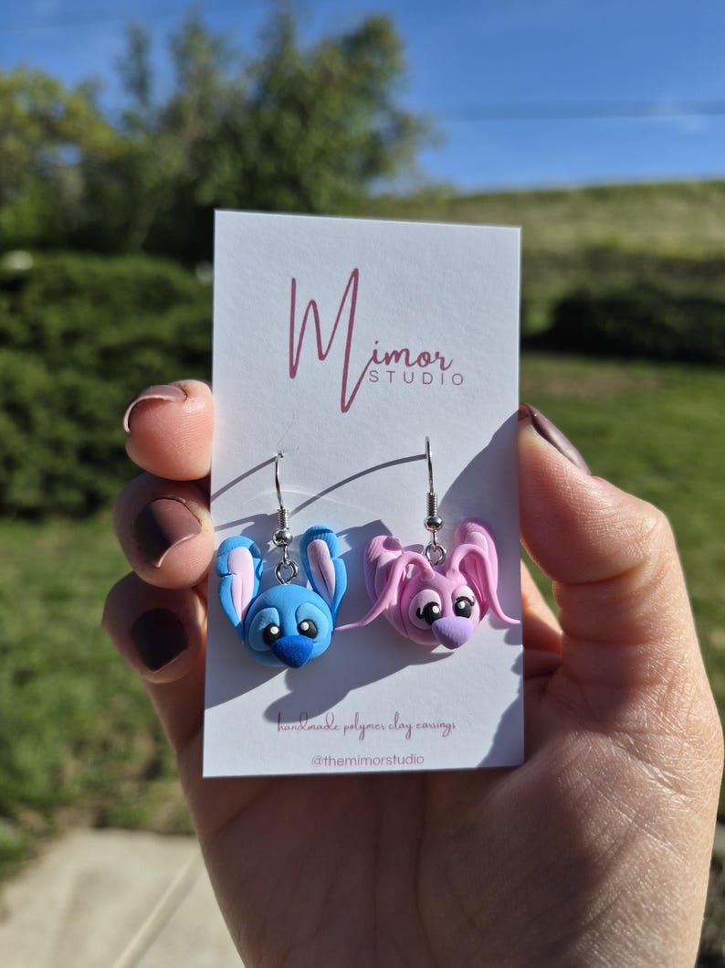 Puede incluir: Pendientes hechos a mano de arcilla polim&eacute;rica, que representan un personaje de dibujos animados azul y otro rosa con orejas grandes. Los pendientes se muestran en una tarjeta blanca con el logotipo "Mimor Studio" y texto. Fondo borroso.