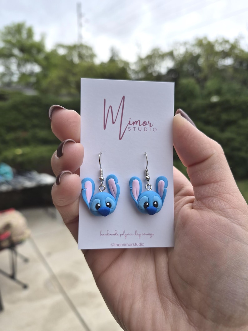 Pu&ograve; includere: Un paio di orecchini in pasta polimerica blu e rosa a forma di personaggio dei cartoni animati. Gli orecchini sono esposti su una carta bianca con la scritta "Mimor Studio" e "handmade polymer clay earrings".