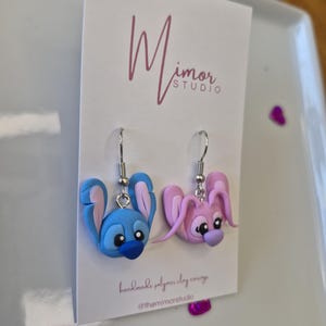 Puede incluir: Un par de pendientes hechos a mano de arcilla polim&eacute;rica con dise&ntilde;os de personajes de dibujos animados. Un pendiente es azul con detalles rosas, el otro es rosa con detalles morados. Los pendientes cuelgan de ganchos plateados. La tarjeta tiene el texto "Mimor Studio".