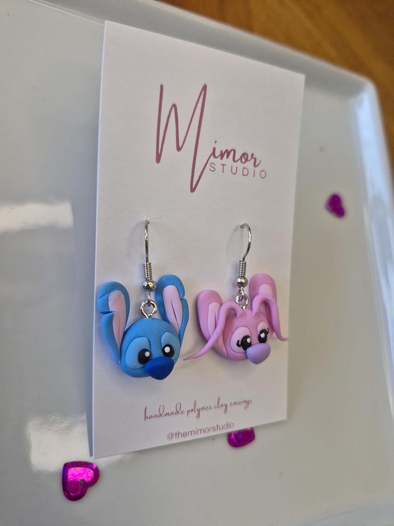Puede incluir: Un par de pendientes hechos a mano de arcilla polim&eacute;rica con dise&ntilde;os de personajes de dibujos animados. Un pendiente es azul con detalles rosas, el otro es rosa con detalles morados. Los pendientes cuelgan de ganchos plateados. La tarjeta tiene el texto "Mimor Studio".