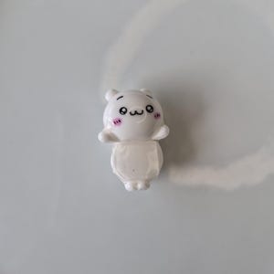 May include: A small, white figurine of a cartoon character with a smiling face, black eyes, and pink blush marks. The figurine has a bear-like head and a rounded body. The background is a plain, light gray surface.