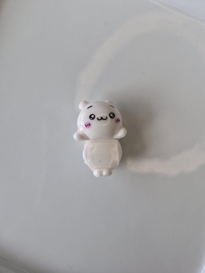 May include: A small, white figurine of a cartoon character with a smiling face, black eyes, and pink blush marks. The figurine has a bear-like head and a rounded body. The background is a plain, light gray surface.