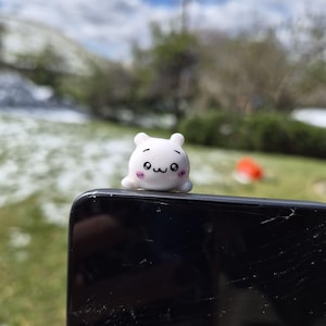 May include: A small, white, cartoon-style figure with a cute face, perched on top of a black smartphone. The figure has black eyes, a small mouth, and pink cheeks. The background shows a blurred outdoor scene with green grass and a blue sky.