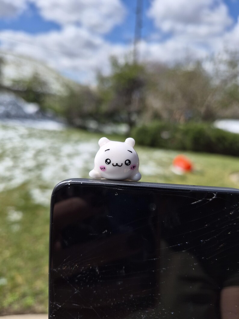 May include: A small, white, cartoon-style figure with a cute face, perched on top of a black smartphone. The figure has black eyes, a small mouth, and pink cheeks. The background shows a blurred outdoor scene with green grass and a blue sky.