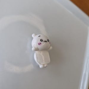 May include: A small, white figurine with a cartoonish design. The figurine has a round head with small ears, a smiling face, and a rectangular body. The figurine is on a white surface.