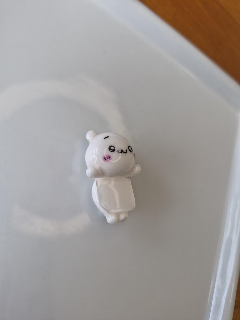 May include: A small, white figurine with a cartoonish design. The figurine has a round head with small ears, a smiling face, and a rectangular body. The figurine is on a white surface.