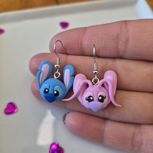 Puede incluir: Un par de pendientes de conejito hechos a mano. Un pendiente es azul con detalles rosas, el otro es rosa con detalles morados. Ambos tienen ganchos plateados. Sobre una superficie blanca con confeti en forma de coraz&oacute;n.