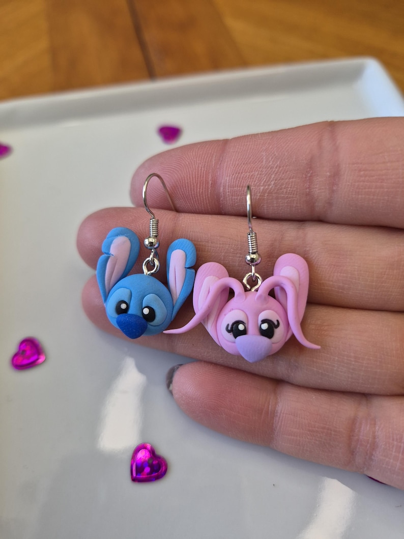 Puede incluir: Un par de pendientes de conejito hechos a mano. Un pendiente es azul con detalles rosas, el otro es rosa con detalles morados. Ambos tienen ganchos plateados. Sobre una superficie blanca con confeti en forma de coraz&oacute;n.