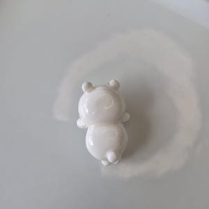May include: A small, white, glossy figurine of a bear-like character. The figurine has rounded features, including a circular body, small arms, and a pair of ears. The bear is standing upright, with its back to the viewer.